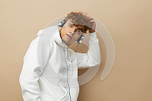 a cute, attractive man stands on a beige background in a white hoodie and listens to music with headphones, scratching