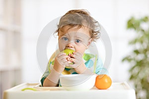 Cute baby 1 years old sitting on high children chair and eating fruits alone in white kitchen