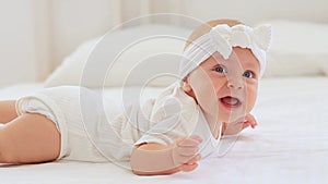 a cute baby with blue eyes of six months is lying on a bed in a white bodysuit in a bright bedroom, a beautiful newborn