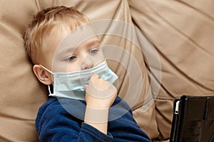 Cute baby boy with blond hair and blue eyes in medical sanitary mask sitting on beige leather sofa with pad digital tablet. Wearin