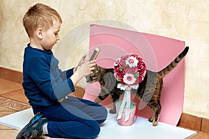 Cute baby boy in blue home wear makes shoot of pretty flowers on pink and white background, with mobile phone.