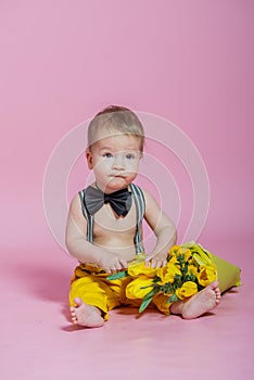 Cute baby boy with bouquet of yellow tulips barefoot in pants in black bow-tie on pink background