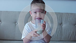 Cute baby boy drinking a glass of Milk sitting on the couch at home. Slow motion little boy drinking water. Close-up