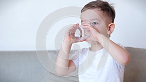 Cute baby boy drinking a glass of water sitting on the couch at home. Slow motion little boy drinking water. Close-up