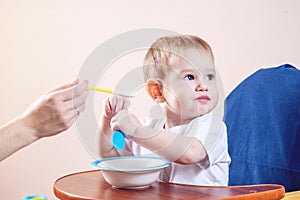 Cute baby boy eating on a chair in the kitchen. Mom feeds holding in hand a spoon of porridge