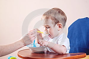 Cute baby boy eating on a chair in the kitchen. Mom feeds holding in hand a spoon of porridge