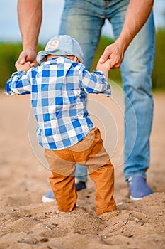 Cute baby boy learns to walk holding daddy`s hands on the beach on the sand