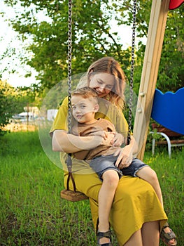 Cute baby boy and mother ride on swing in playground on green lawn. Summertime photo for ad or blog about motherhood