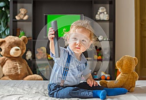 Cute baby boy playing with the remote control to watch TV sitting on a couch with his teddy bear, at home