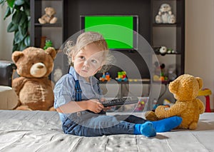 Cute baby boy playing with the remote control to watch TV sitting on a couch with his teddy bear, at home