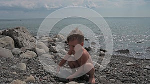 Cute baby boy plays with stones on the beach, on the seashore and climbs on the rocks. Summer holidays and sea holidays