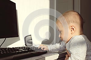 A cute baby boy is sitting at the computer and looks attentively with admiration