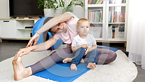 Cute baby boy sitting on fitness mat and looking at his mother stretching legs before sports training. Concept of healthcare,