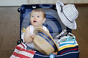 Cute baby boy sitting in the suitcase with hat in his hands, packed for vacation