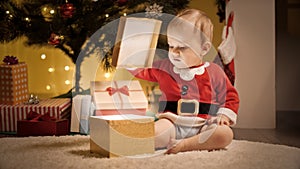 Cute baby boy sitting under Christmas tree and opening lid of golden gift box