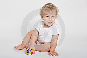 Cute baby boy toddler playing with toy car indoor, posing isolated over light background, wearing white bodysuit, looking directly