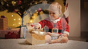 Cute baby boy unwrapping and looking inside Christmas present box under decorated Christmas tree. Families and children