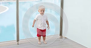 A cute baby with candy runs on the summer terrace with a pool in the background.