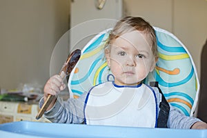 Cute baby child getting messy eating cereals or porridge by itself, with a wooden spoon