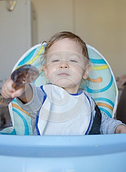 Cute baby child getting messy eating cereals or porridge by itself, with a wooden spoon