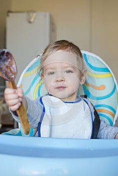 Cute baby child getting messy eating cereals or porridge by itself, with a wooden spoon