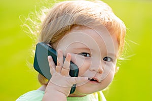 Cute baby face closeup on green grass in summertime. Funny little kid portrait with mobile phone, smartphone on nature