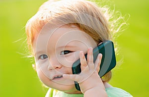 Cute baby face closeup on green grass in summertime. Funny little kid portrait with mobile phone, smartphone on nature