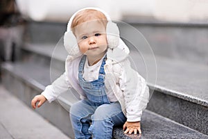 Cute baby girl 1-2 year old wear fluffy white headphones, denim suit pants and knitted sweater posing on city street outdoors.