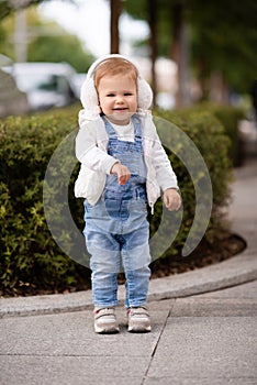 Cute baby girl 1-2 year old wear fluffy white headphones, denim suit pants and knitted sweater posing on city street outdoors.