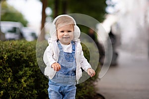 Cute baby girl 1-2 year old wear fluffy white headphones, denim suit pants and knitted sweater posing on city street outdoors.