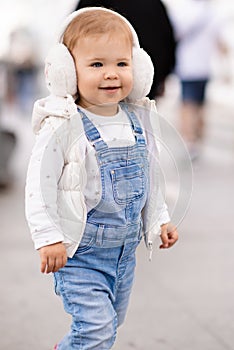 Cute baby girl 1-2 year old wear fluffy white headphones, denim suit pants and knitted sweater posing on city street outdoors.