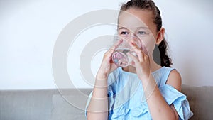 Cute baby girl drinking a glass of water sitting on the couch at home. Slow motion little boy drinking water. Close-up