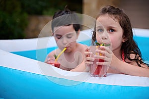 Cute baby girl drinks cocktail while having fun with her older brother in outdoor swimming pool at house backyard garden