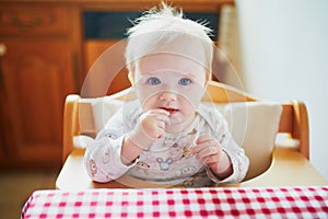 Cute baby girl feeding herself with finger food in the kitchen