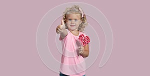 Cute baby girl holding a lollipop smiles and shows thumb up isolated over pink background