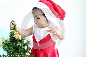 Cute baby girl kid in Santa Claus red costume trying to decorate Christmas tree in white living room, beautiful little child