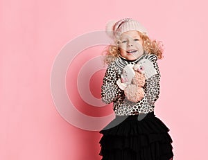 Cute baby girl in knitted hat and gloves clutched something to her chest or shivering cold on pink