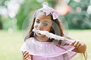 Cute baby girl in pink dress eats white cotton candy in the park in summer