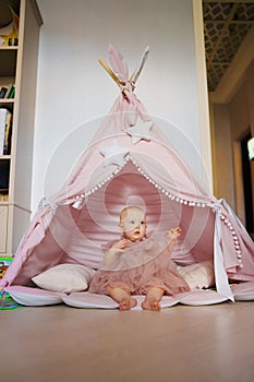 Cute baby girl in pink dress sitting in teepee tent for children& x27;s room.