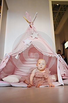 Cute baby girl in pink dress sitting in teepee tent for children& x27;s room.