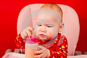 Cute baby girl playing with her food on the red background. Little kid infant with a bottle of milk on a red background.