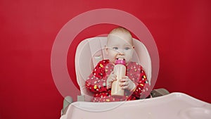 Cute baby girl playing with her food on the red background. Little kid infant with a bottle of milk on a red background.
