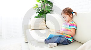 Cute baby girl reading book at home sitting on sofa