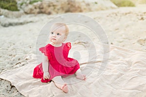 Cute baby girl in a red dress sitting and playing on the beach
