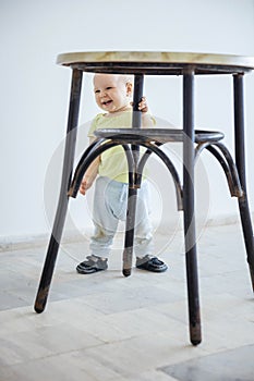 Cute baby girl standing while gripping leg of stool and laughing