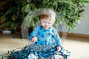 Cute baby girl taking down holiday decorations from Christmas tree. child holding light garland.