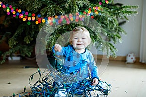 Cute baby girl taking down holiday decorations from Christmas tree. child holding light garland. Family after