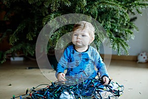 Cute baby girl taking down holiday decorations from Christmas tree. child holding light garland. Family after