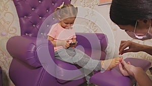 Cute baby girl using smart phone at pedicure procedure at beauty spa salon.
