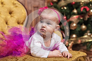 Cute baby girl wearing pink skirt and red headband, laying on couch in front of christmas tree.